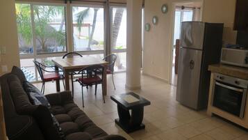 A living room with a sofa, a dining table with chairs, a kitchen area with a refrigerator and microwave, and large windows with a view of palm trees.