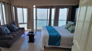 Una habitación con una cama, un sofá y una mesita. Hay ventanas amplias con cortinas, y se aprecia vista al mar.