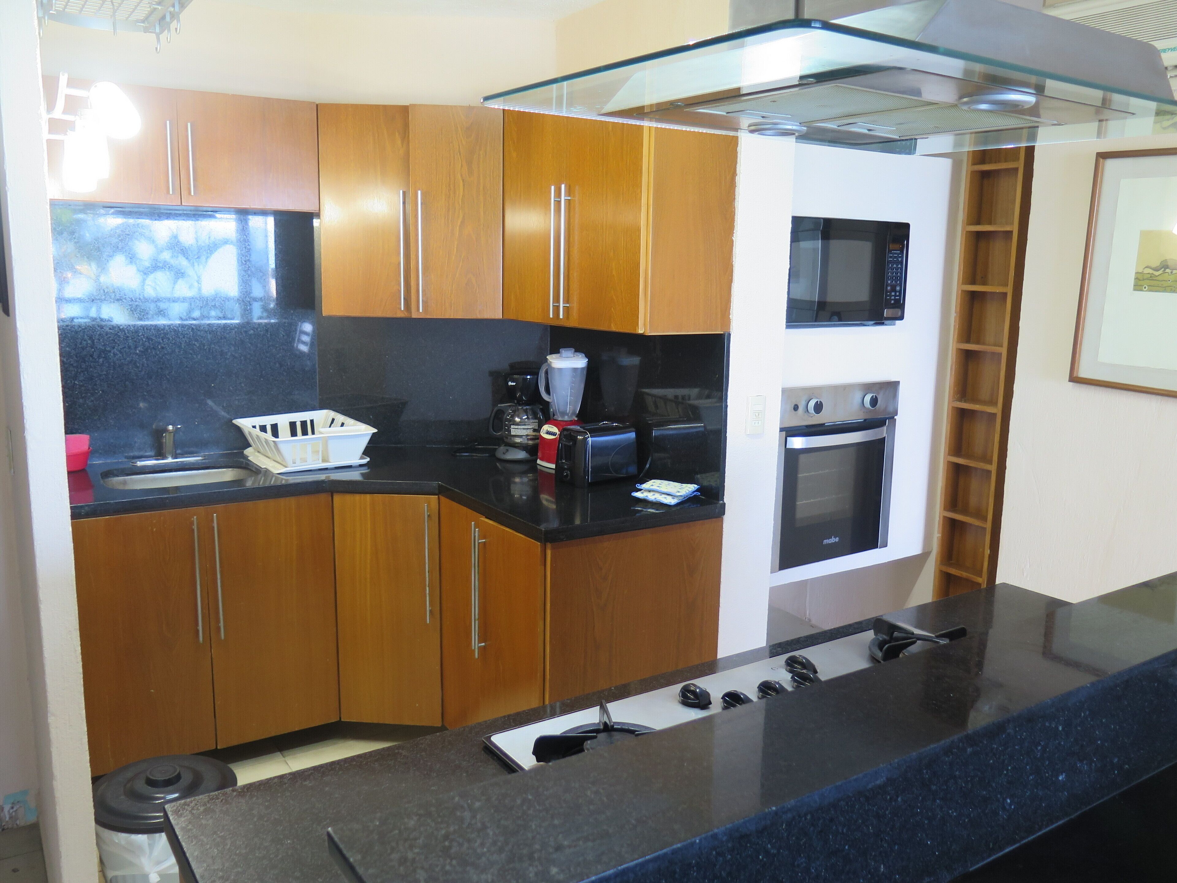 A modern kitchen with wooden cabinets, a black countertop, a built-in microwave, an oven, and a glass shelf above the counter.