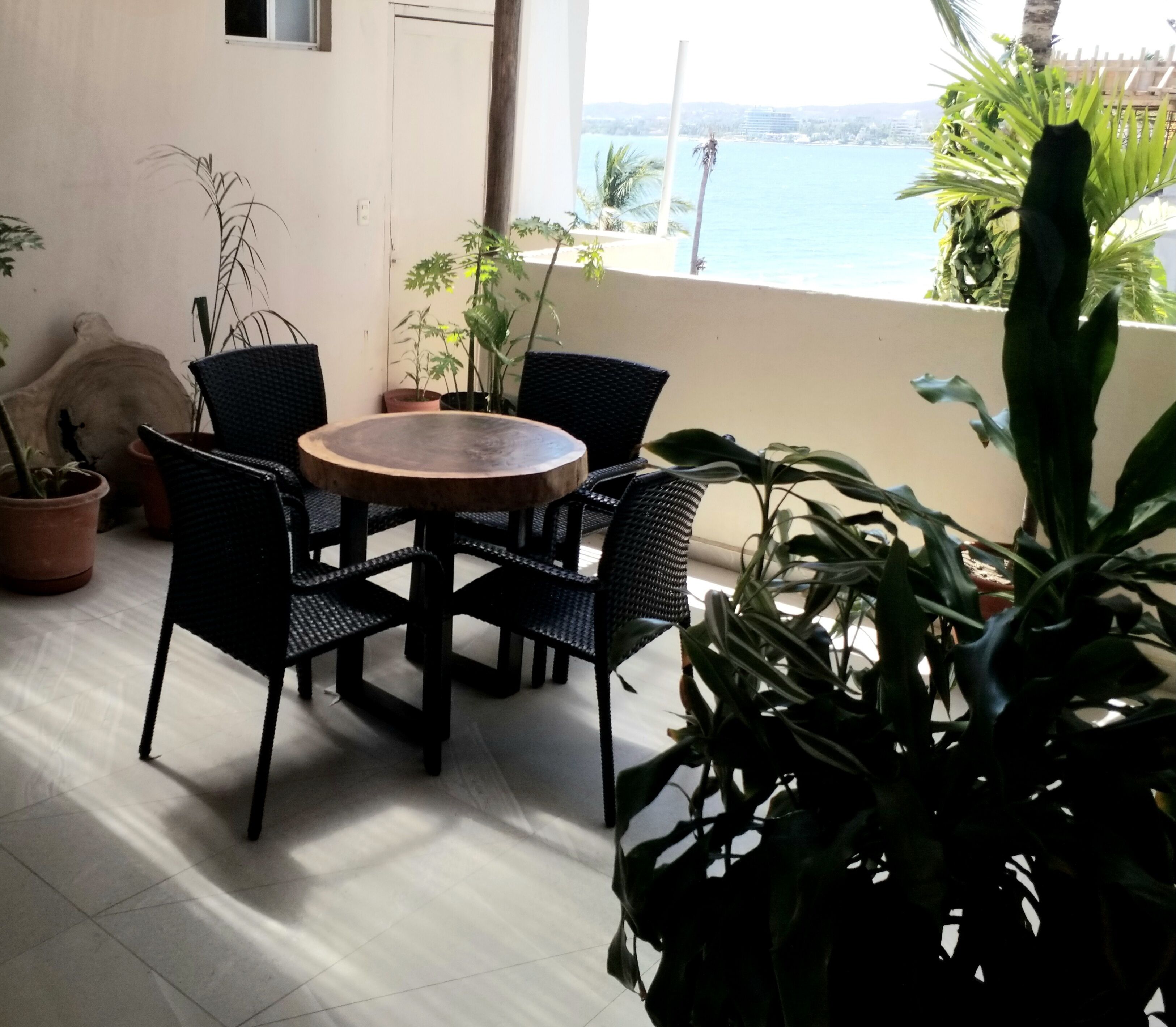 Une table ronde en bois, entourée de quatre chaises en osier noir, et agrémentée de plantes en pot, sur un balcon offrant une vue sur la mer.