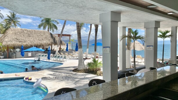 Zona de piscina con sillas de descanso y sombrillas, una estructura de techo de paja y vistas al mar.