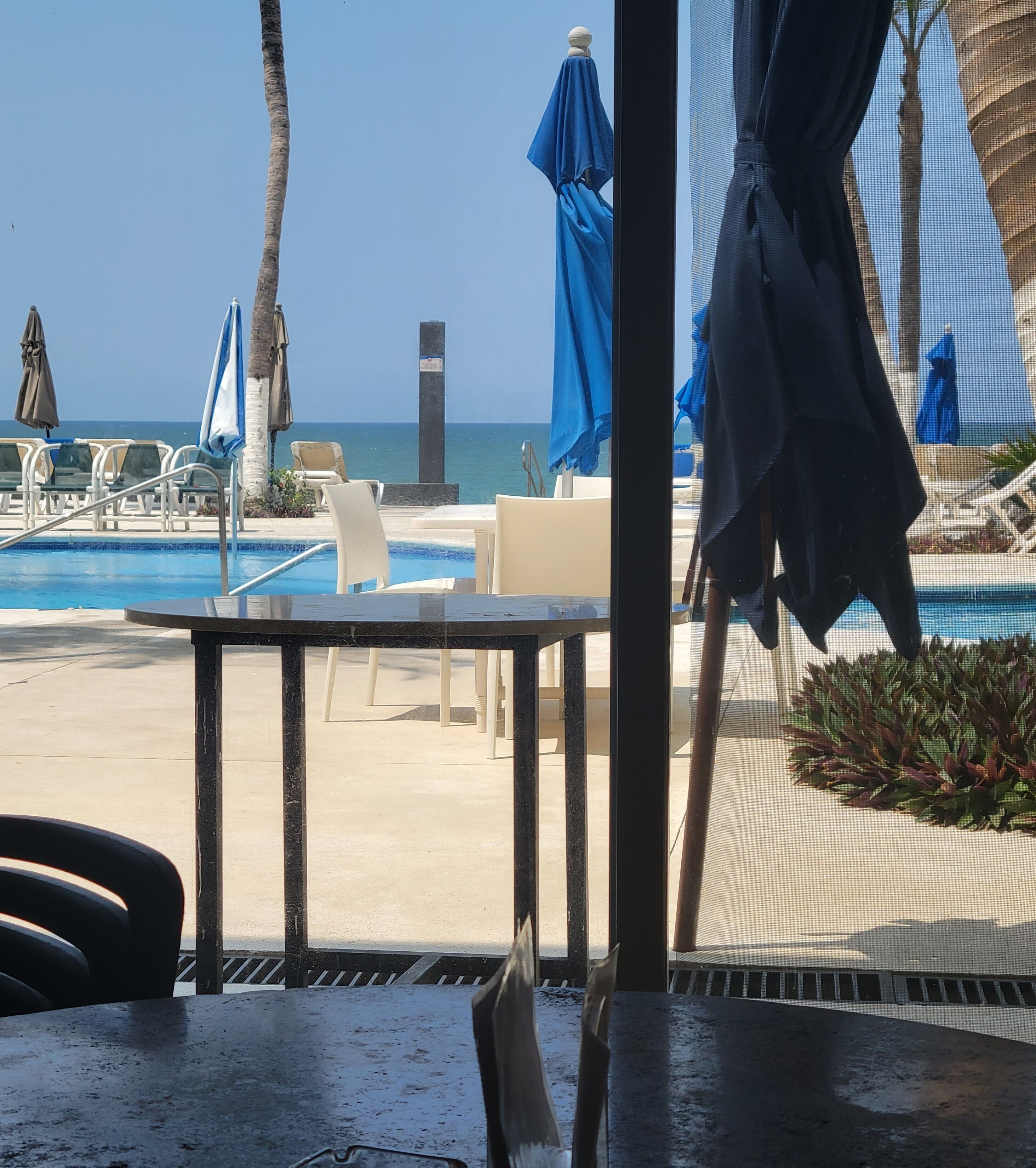 A poolside area with lounge chairs and umbrellas, a table, and a view of the ocean.