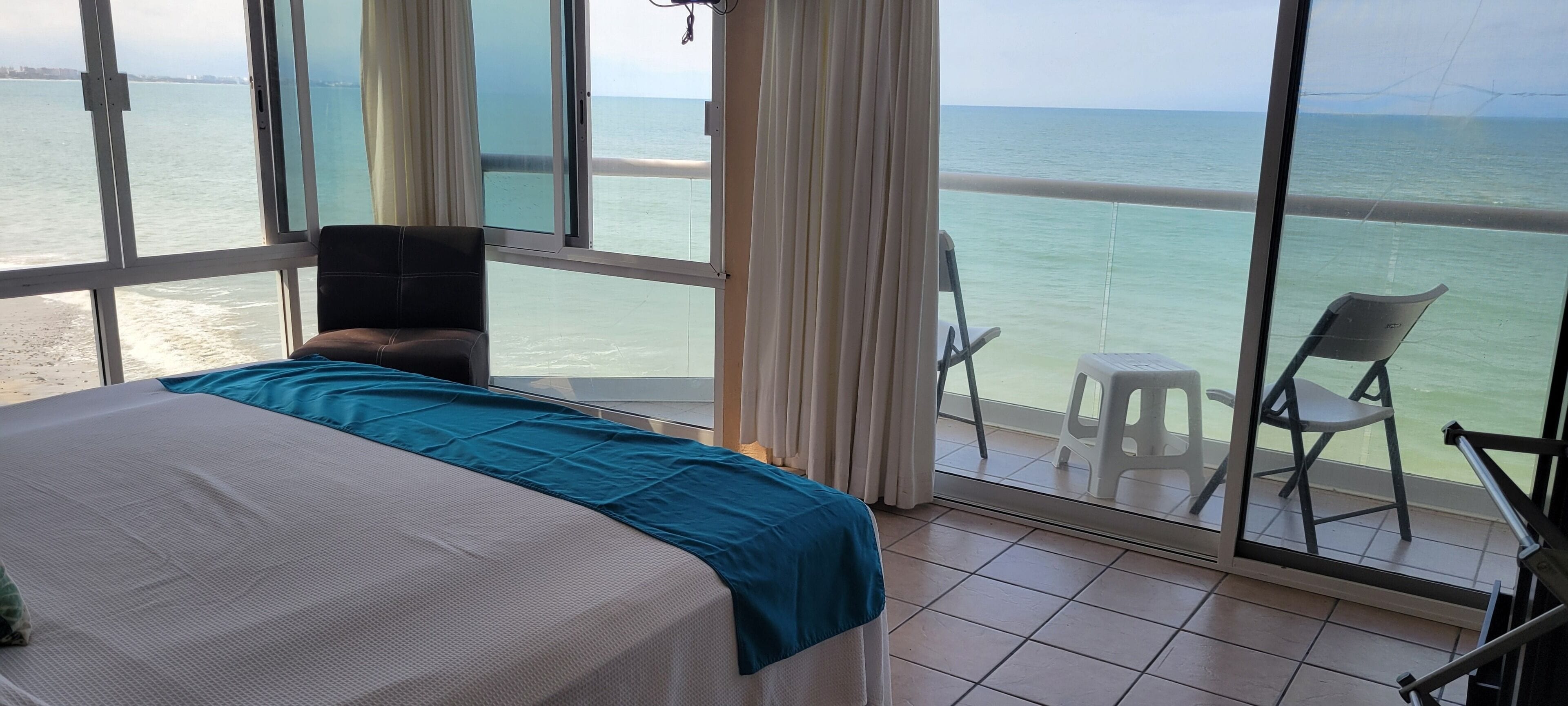A room with a bed, a balcony with chairs, and a view of the sea.