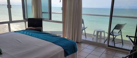 A room with a bed, a balcony with chairs, and a view of the sea.
