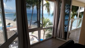 Habitación con ventanas amplias que dan a una playa y al océano, con piso de baldosas y un sofá negro.
