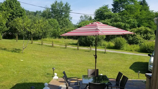 An outdoor patio area with a table and chairs, a red and white striped umbrella, and a view of a grassy field and trees in the background. - Catskills Modern Comfort Home Mountainous Backdrop (East Branch)