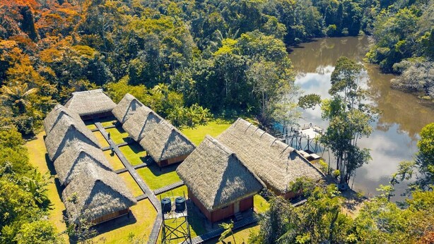 A traditional thatched-roof building complex with a central courtyard, surrounded by dense forest and a body of water.