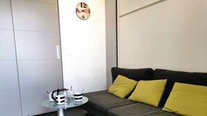 Een moderne woonkamer met een grijze bank, gele kussens, een glazen salontafel en een wandklok.
