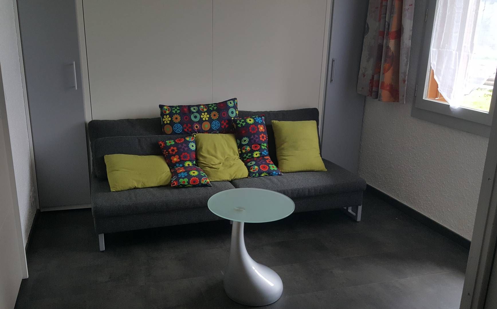 Ein graues Sofa mit bunten, blumigen Kissen, ein kleiner runder Tisch und ein Fenster mit Vorhängen.