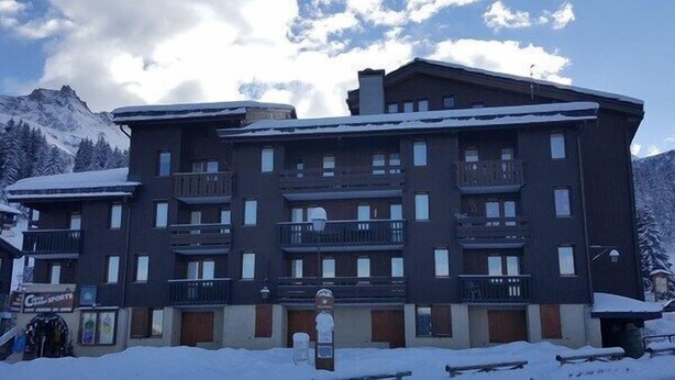 Een hotel met meerdere verdiepingen, balkons, een bord met 'skigebied' en besneeuwde omgeving.