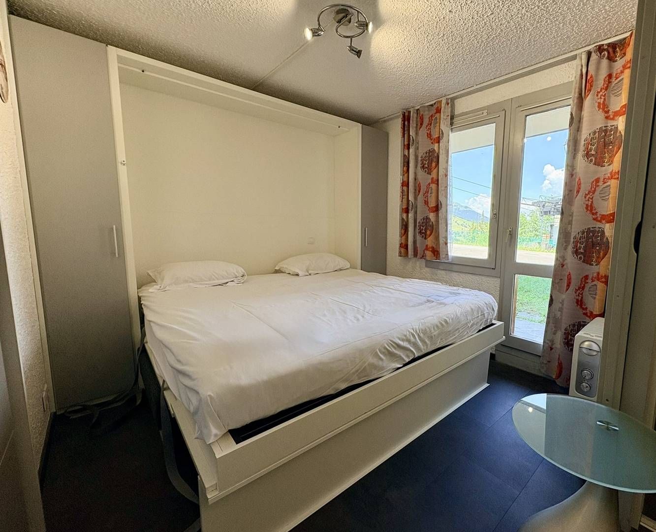 A single-bed room with a built-in wardrobe, a small table, and a window with curtains.
