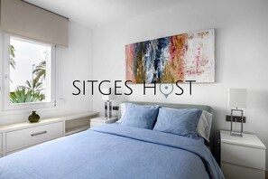 A modern bedroom with a large bed, a window with a view of palm trees, a colorful abstract painting on the wall, and a nightstand with a lamp.