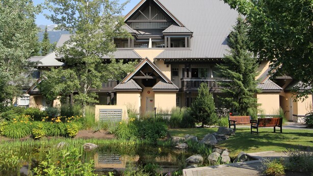A two-story building with a symmetrical roof design, surrounded by a landscaped garden with a pond and benches. - Lagoons Condos by Whistler Retreats (Whistler)