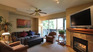 A living room with a fireplace, a television, a ceiling fan, and a large window. - Lagoons Condos by Whistler Retreats (Whistler)