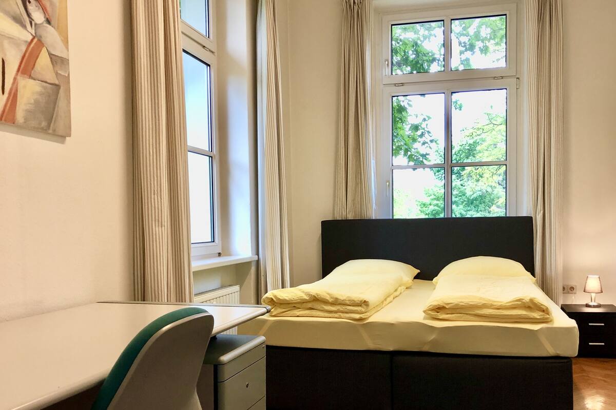 A bedroom with a bed, a desk, and a chair. There are curtains on the windows, and a lamp on the bedside table.