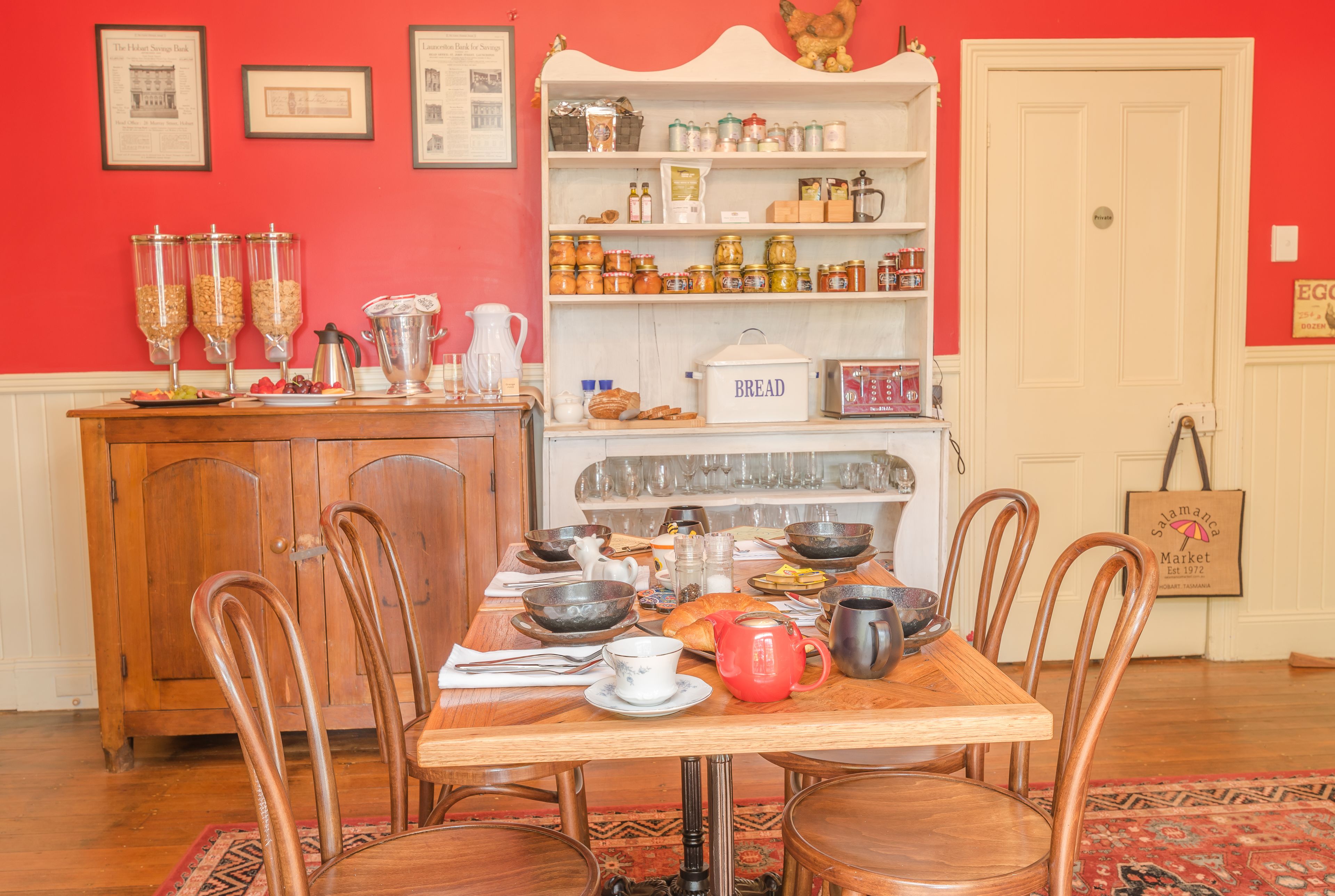 A dining area with a wooden table set for six, surrounded by wooden chairs. A white cabinet with glass jars and a bread box is to the left.