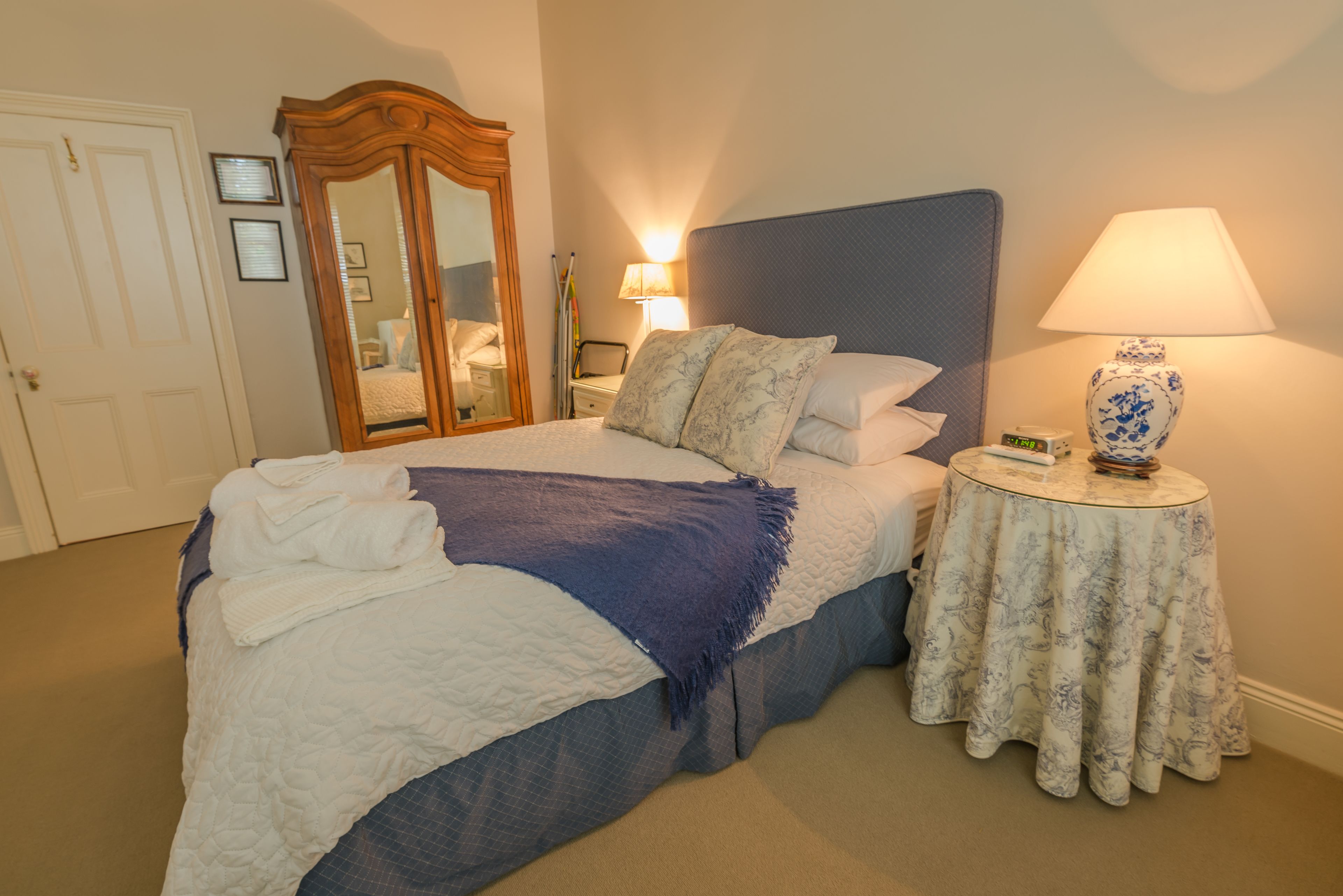 A neatly made bed with a blue blanket and pillows, a bedside table with a lamp and a vase, and a mirror on the wall.
