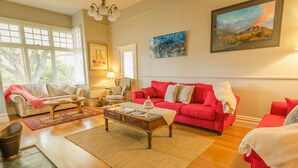 Een woonkamer met een rode bank, een beige bank, een houten salontafel en een rood vloerkleed. Er hangen schilderijen aan de muur en er is een raam met zonwering.