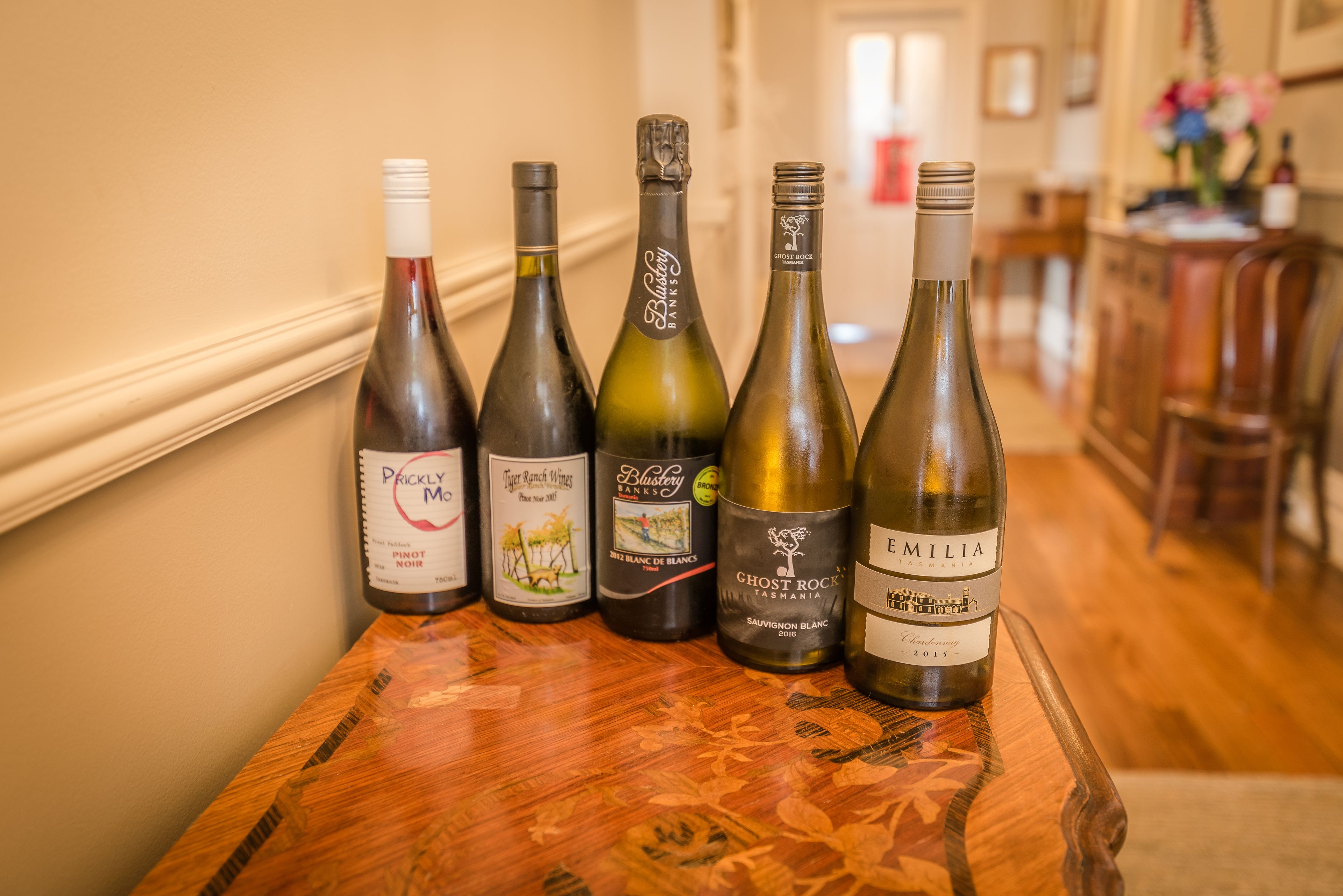 Une rangée de bouteilles de vin est posée sur une table en bois, dans une pièce dont le sol est également en bois et dont le mur est blanc.