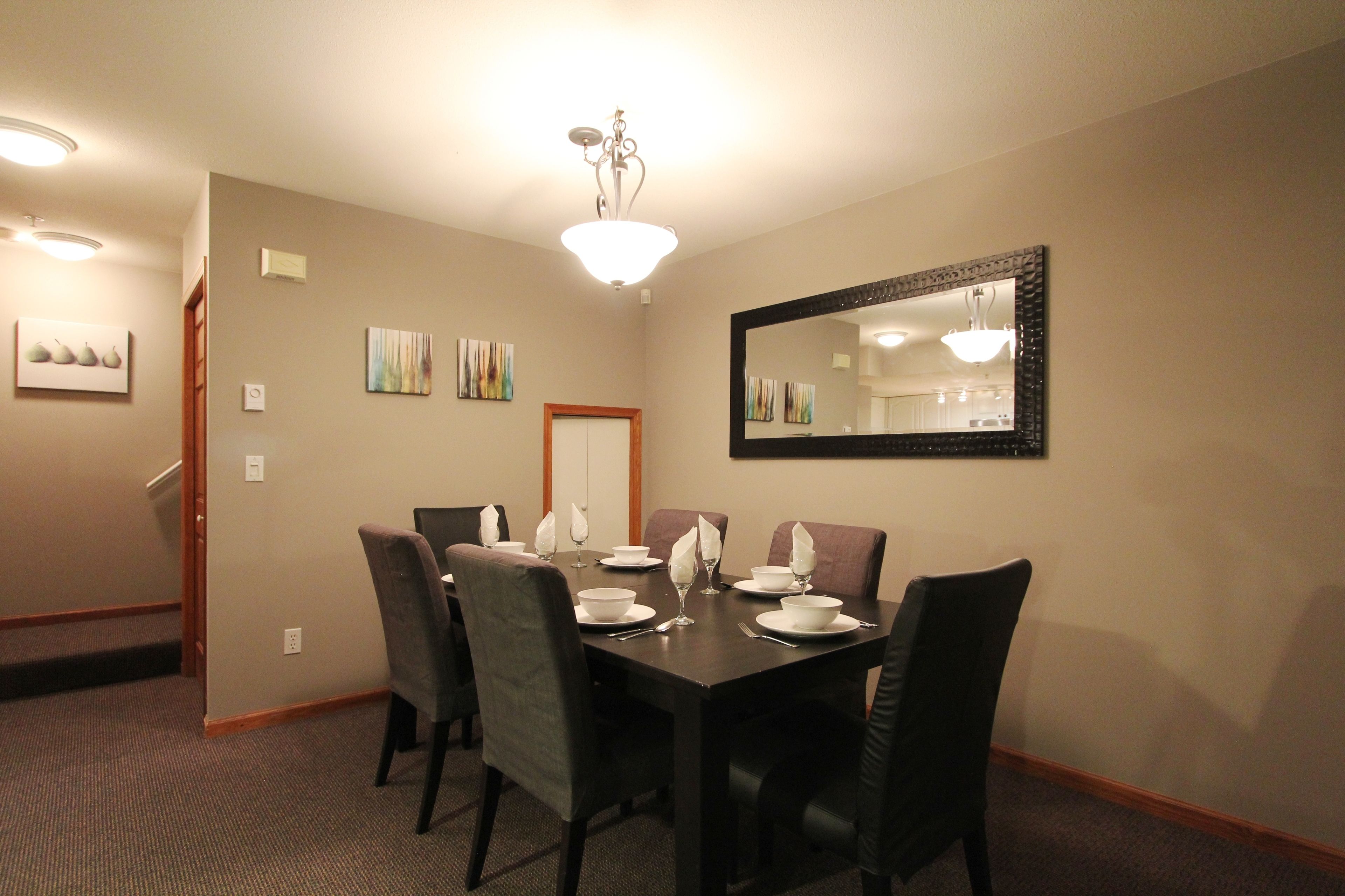 A dining area with a dark wooden table set for four, surrounded by chairs, with a mirror on the wall and artwork above the table.