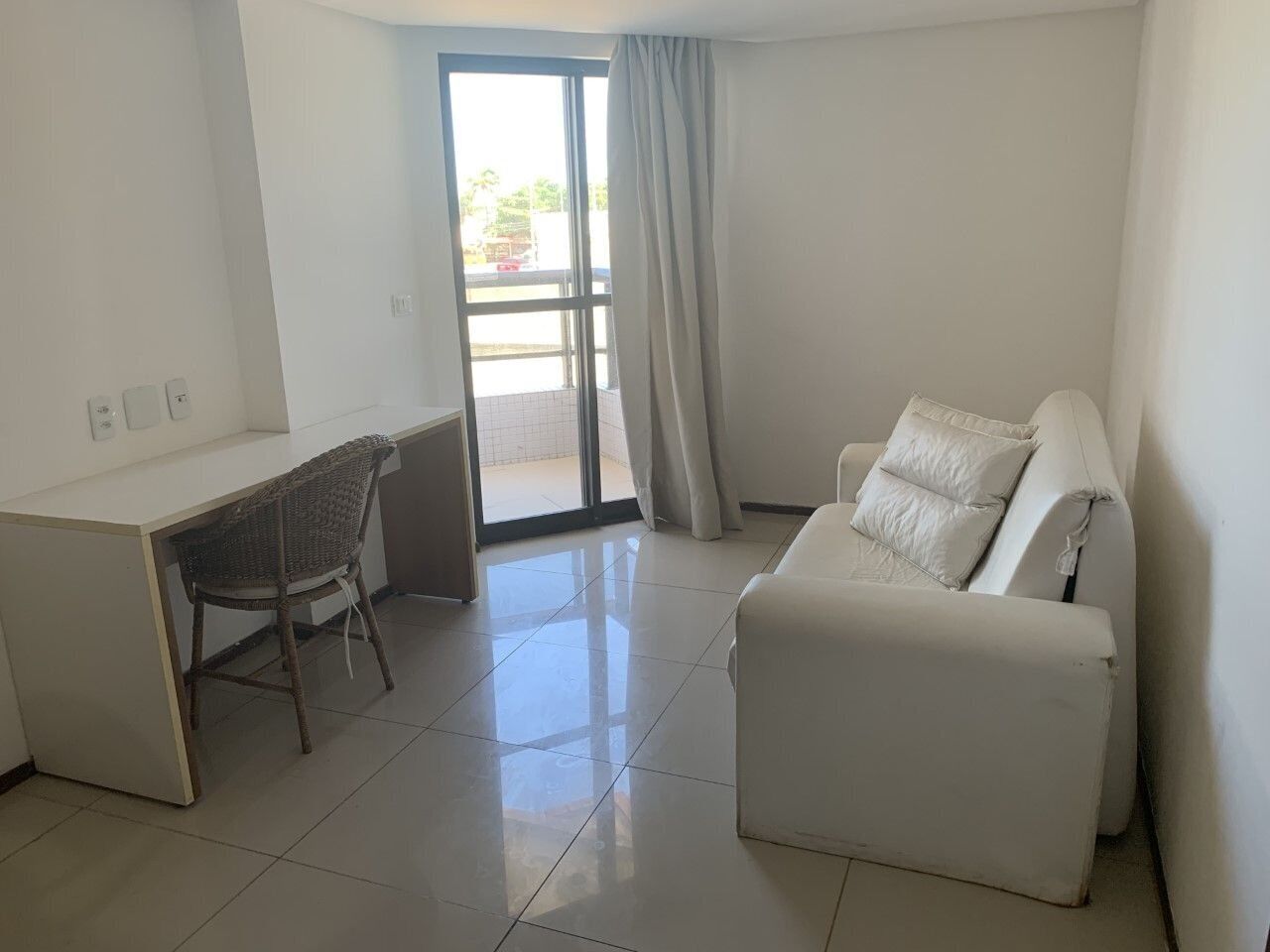 Una habitación con un escritorio blanco, una silla de mimbre, un sofá blanco y un ventanal corredero que da a un espacio exterior.