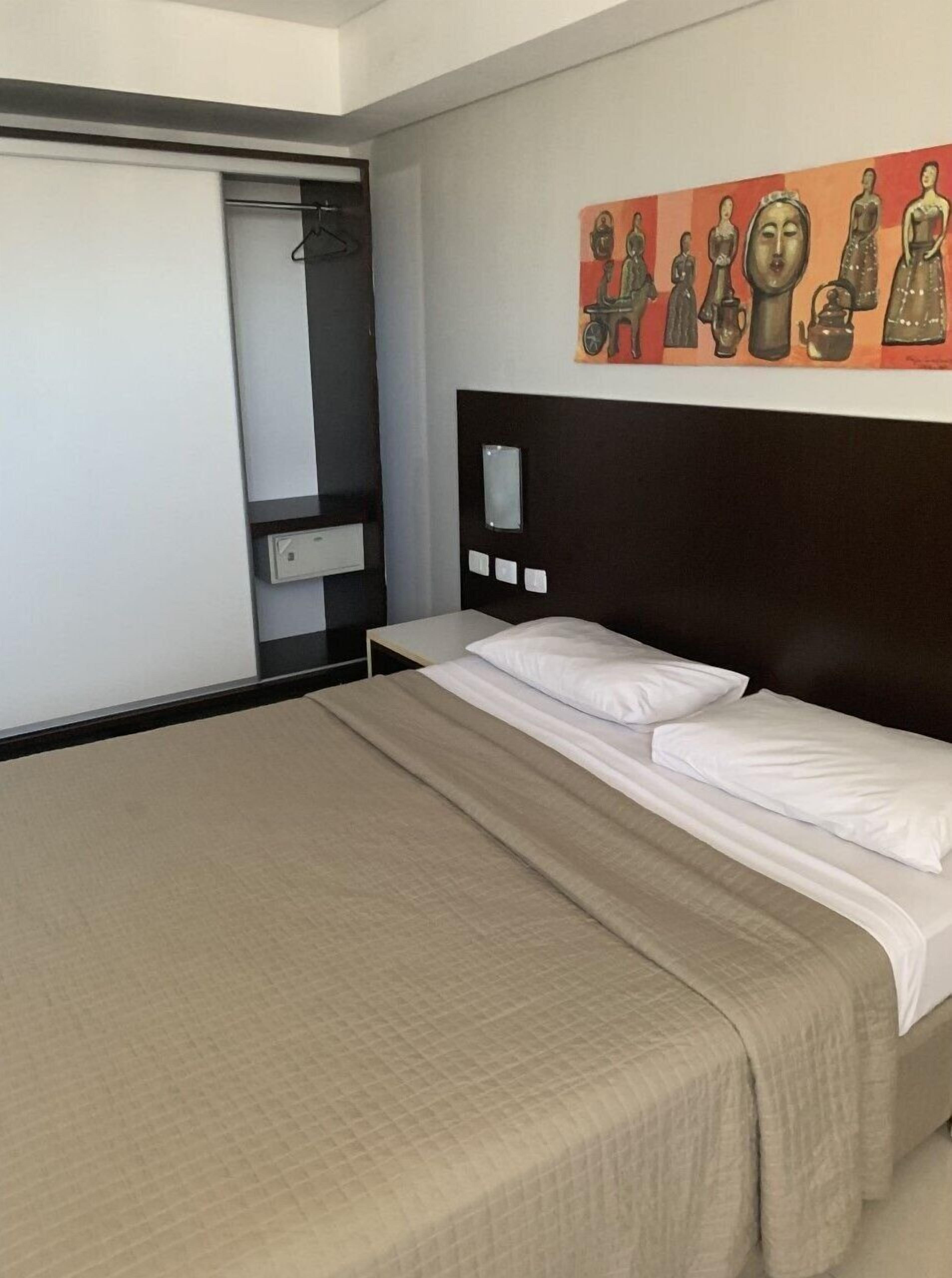 Hotellihuone, jossa on sänky, vaatekaappi, yöpöytä ja seinään kiinnitetty valaisin.