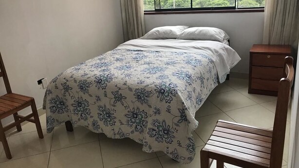 A bedroom with a bed, two wooden chairs, a nightstand, and a window with curtains. - Hospedaje Las Flores (Trujillo)