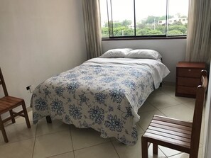 A bedroom with a bed, two wooden chairs, a nightstand, and a window with curtains.