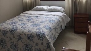 A bedroom with a bed, a wooden nightstand, a chair, and a window with curtains. - Hospedaje Las Flores (Trujillo)