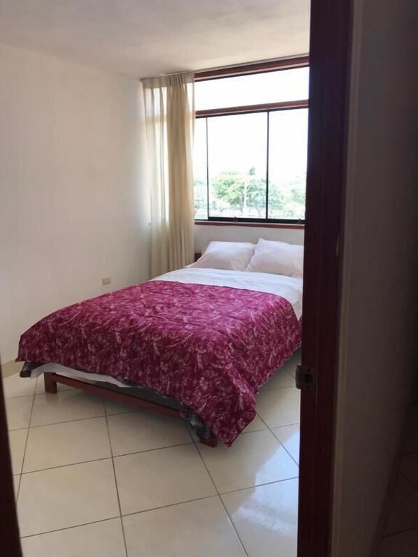 A bedroom with a bed, a window with curtains, and a tiled floor.