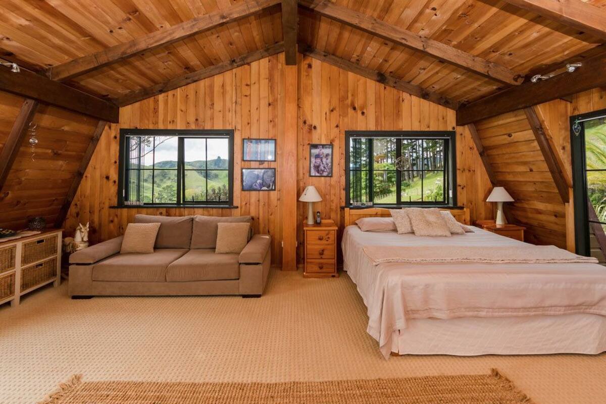 A cozy room with a wooden ceiling, a bed, a sofa, and two windows offering a view of greenery.