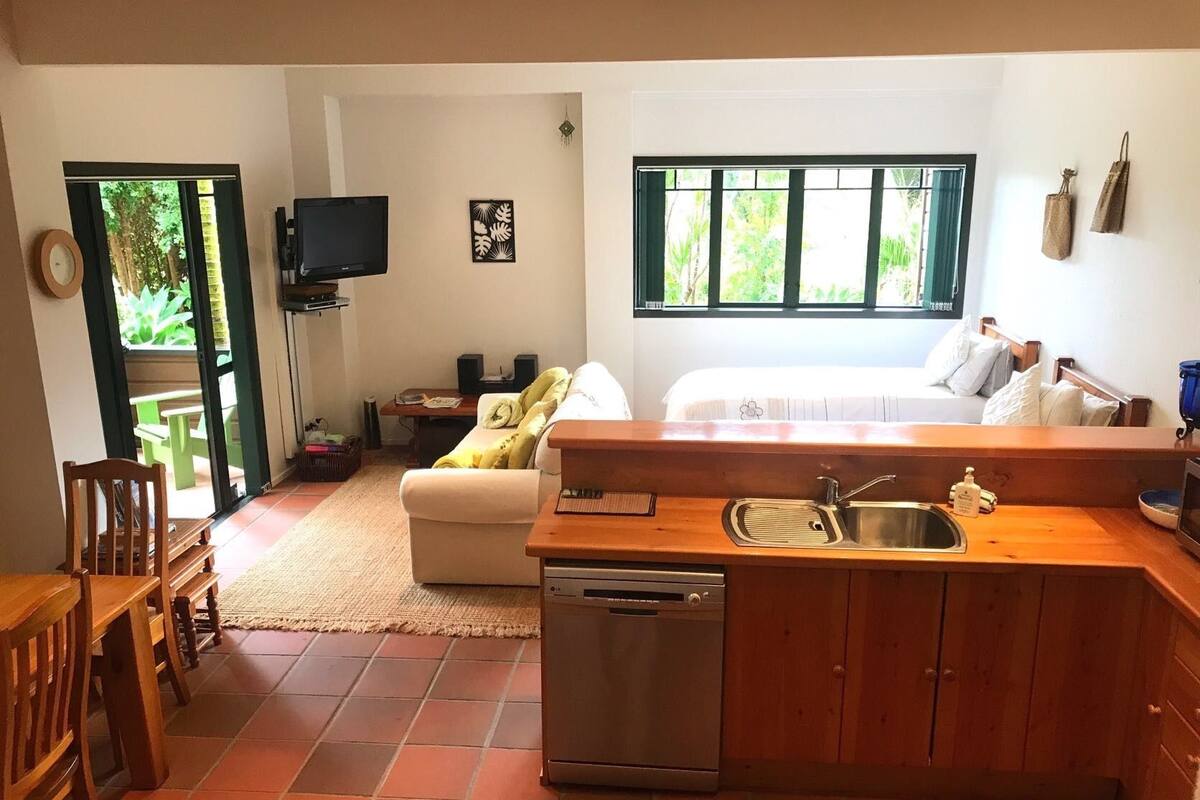 A compact kitchen with a sink, wooden cabinets, and a dishwasher. There is a dining area with a table and chairs, a sofa, and a television mounted on the wall.