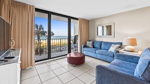 Uma sala de estar moderna com um sofá azul, um banquinho vermelho e uma mesa de vidro. Uma janela ampla oferece vista para a praia e palmeiras.