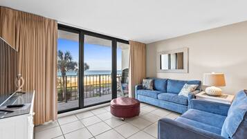 Un salon moderne avec un canapé bleu, un pouf rouge et une table en verre. Une grande fenêtre offre une vue sur la plage et les palmiers.