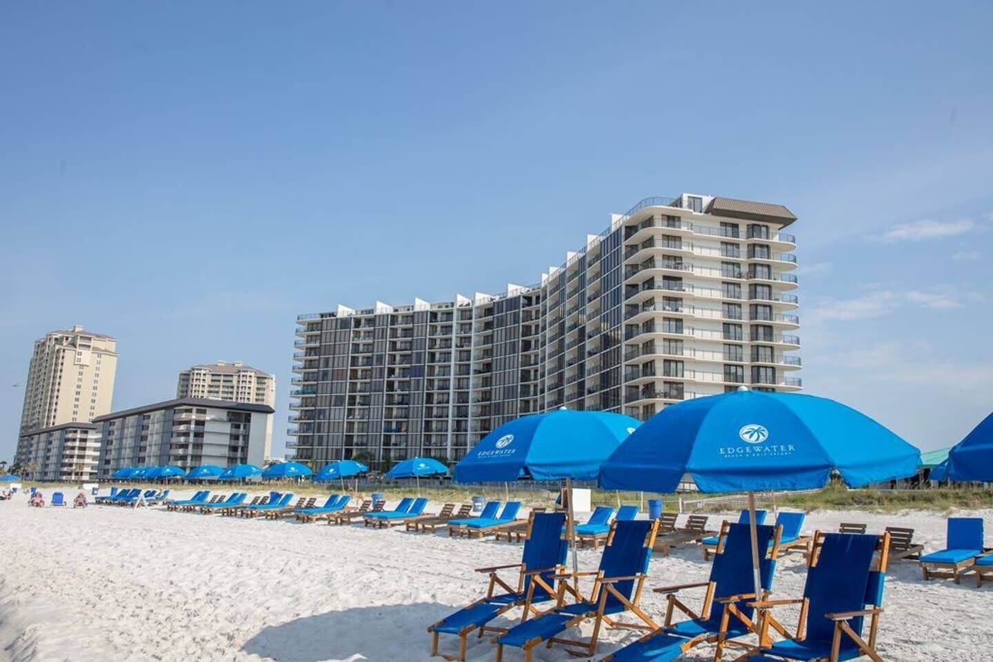 Un hôtel situé en bord de mer, avec plusieurs étages, des parasols bleus et des chaises de plage.