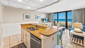 Cozinha com bancadas de granito, máquina de lavar louça e área de jantar com mesa e cadeiras. Há duas imagens emolduradas na parede, e uma grande janela com vista para o oceano.