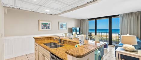 A kitchen with granite countertops, a dishwasher, and a dining area with a table and chairs. There are two framed pictures on the wall, and a large window with a view of the ocean.