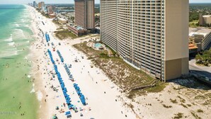 Un hotel frente a la playa con varios edificios altos, una playa de arena con hileras de sombrillas y una vista despejada del océano.