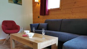 Une table en bois, une chaise rouge et un canapé gris se trouvent dans une pièce aux murs en bois, avec une fenêtre ornée de rideaux.