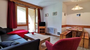 Une pièce chaleureuse avec un canapé, une petite table, un fauteuil rouge, un téléviseur et un coin repas avec une table mise pour deux personnes.