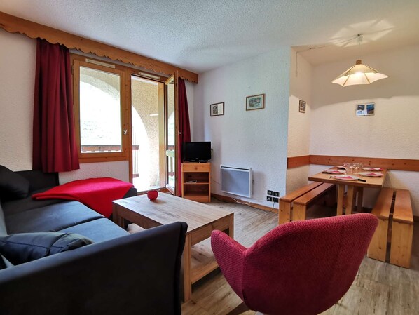 Una habitación acogedora con sofá, una mesita, una silla roja, televisor y un comedor con una mesa dispuesta para dos.