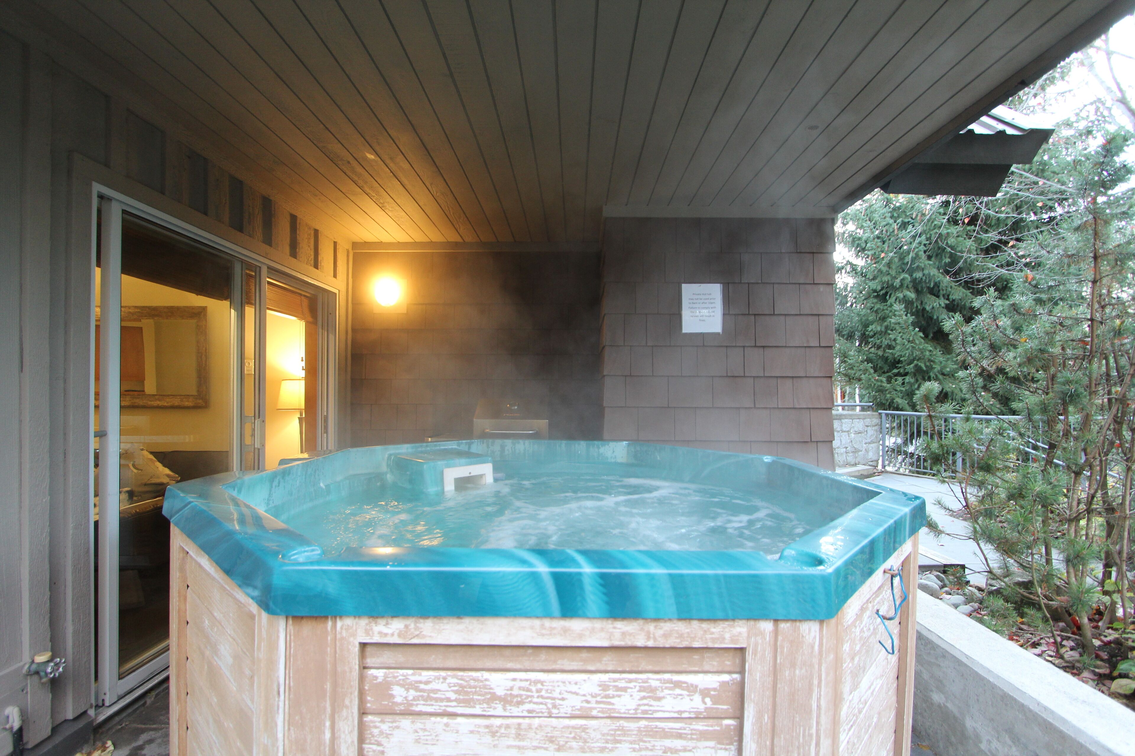 A hot tub with a wooden exterior and a glass top, located under a covered patio area with a brick wall and a sign on the wall.