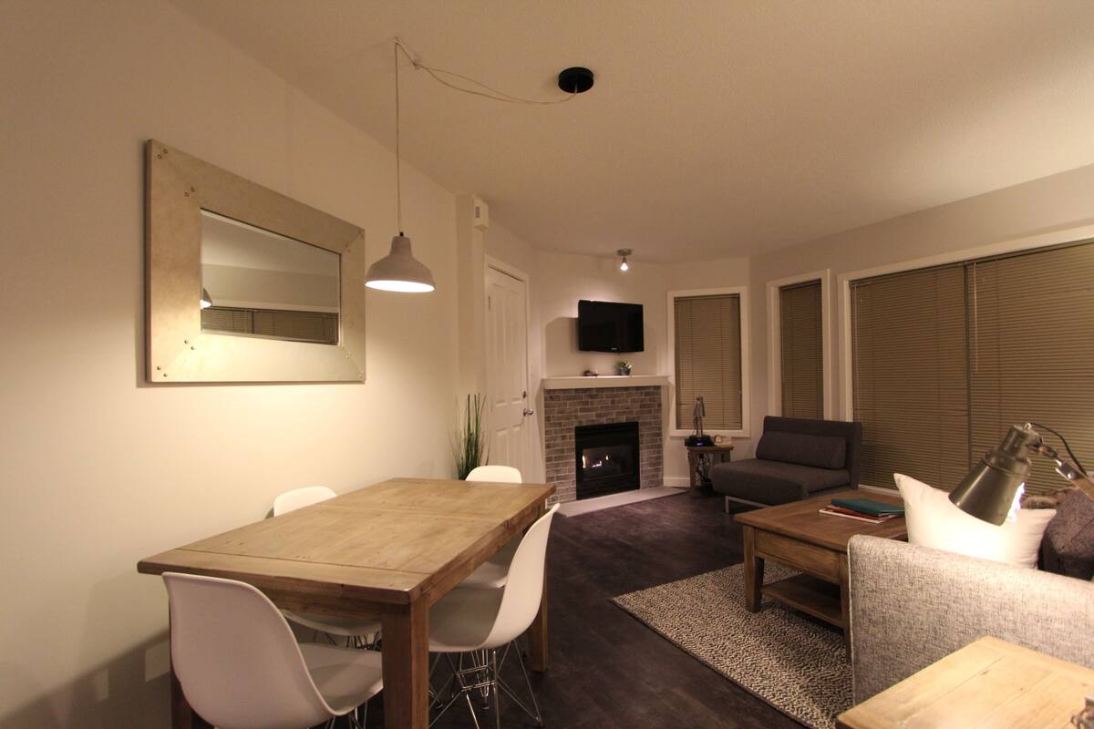 Ein Wohnzimmer mit Kamin, einem darüber montierten Fernseher und einem Essbereich mit Holztisch und weißen Stühlen.