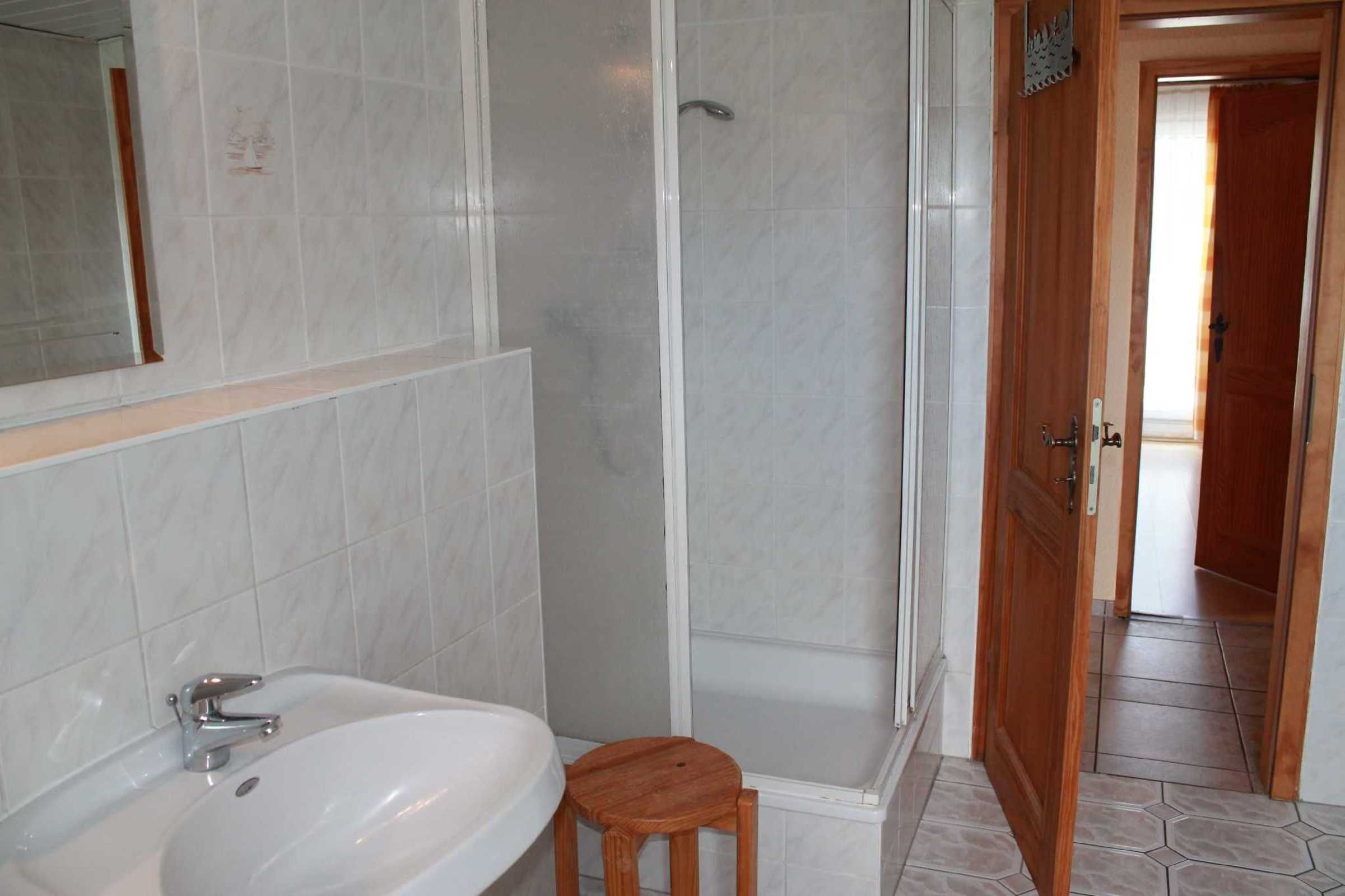 A bathroom with a white sink, a wooden stool, and a glass shower enclosure.