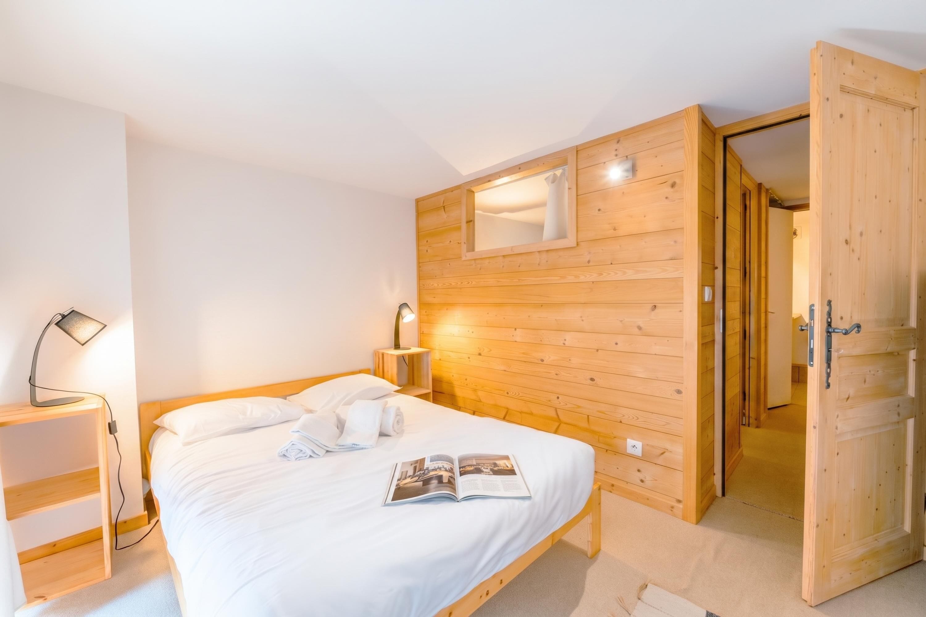 Une chambre à coucher avec une tête de lit en bois, un lit recouvert de draps blancs, une lampe de chevet et un magazine posé sur le lit.
