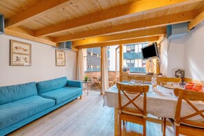 Een kamer met een blauwe bank, houten balken in het plafond, een eettafel met stoelen en een aan de muur bevestigde televisie.