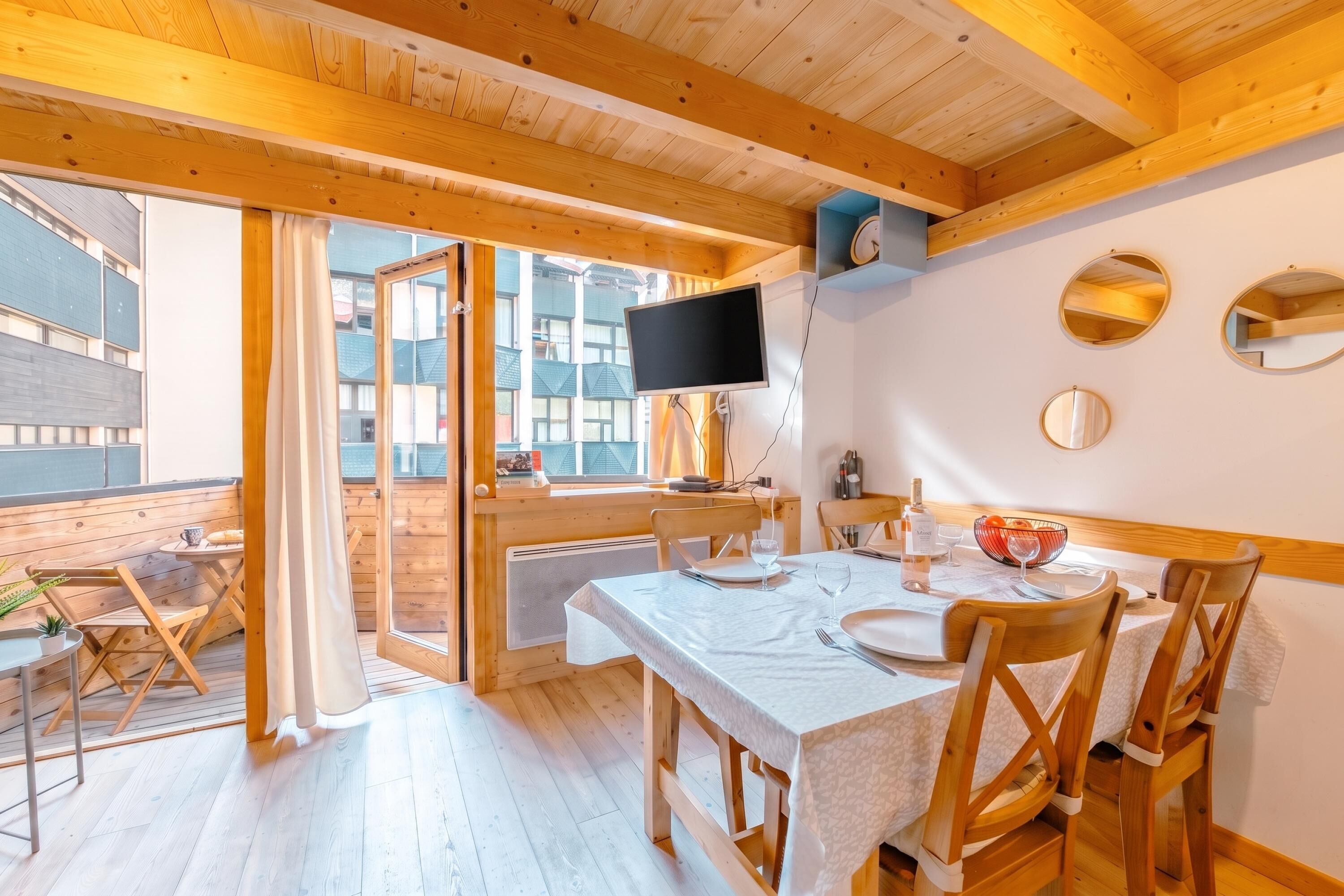 A wooden interior with a dining area, a TV mounted on the wall, and a view through the glass door.