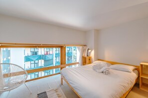 Une chambre à coucher avec un lit, une table de chevet, une lampe, une chaise et une vue sur un immeuble par la fenêtre.