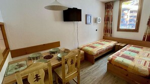 Una habitación con una mesa de comedor de madera dispuesta para dos personas, dos camas individuales con ropa de cama estampada, un televisor montado en la pared y una ventana con cortinas.