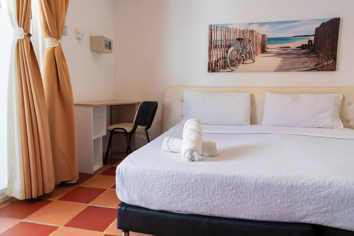 A hotel room with a bed, a desk, a chair, a painting on the wall, and a window with curtains.